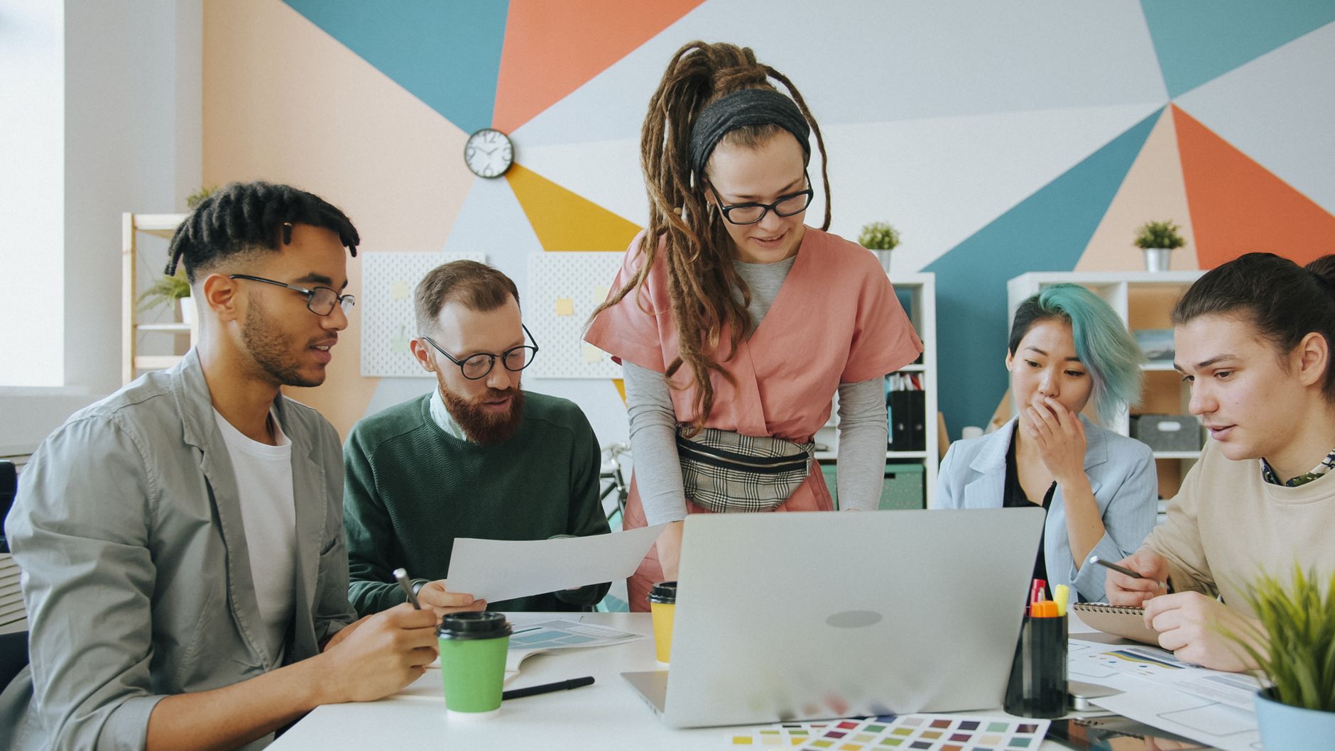 Diverse team of young adults collaborating around a laptop in a colorful modern office setting.